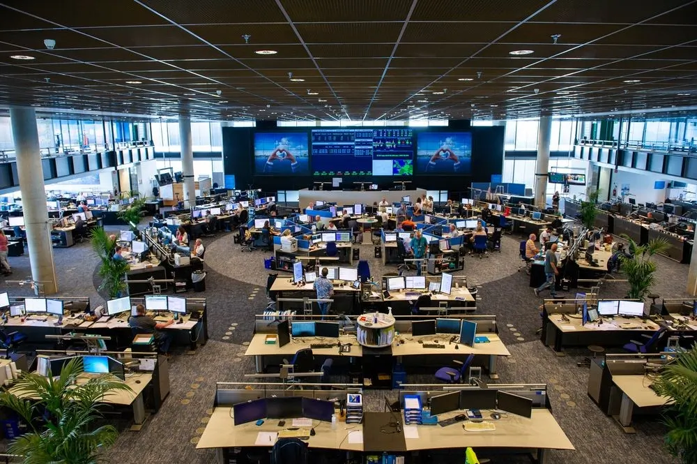 KLM Operations Control Centre at Schiphol, an open-plan room of staffed workstations beneath a live video wall of flight data.