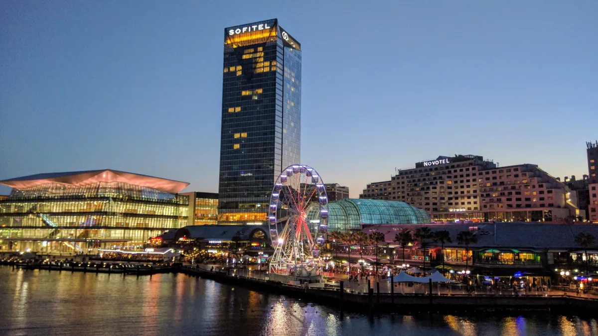 The Sofitel and Novotel towers above Darling Harbour in Sydney at dusk, with the waterfront promenade and ferris wheel lit below