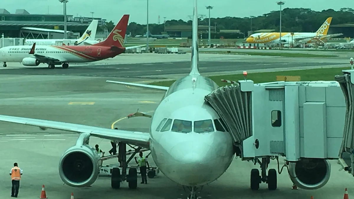 A busy apron at Singapore Changi Airport with an unmarked white Airbus A320 at a jet bridge in the foreground and aircraft from two other carriers behind it