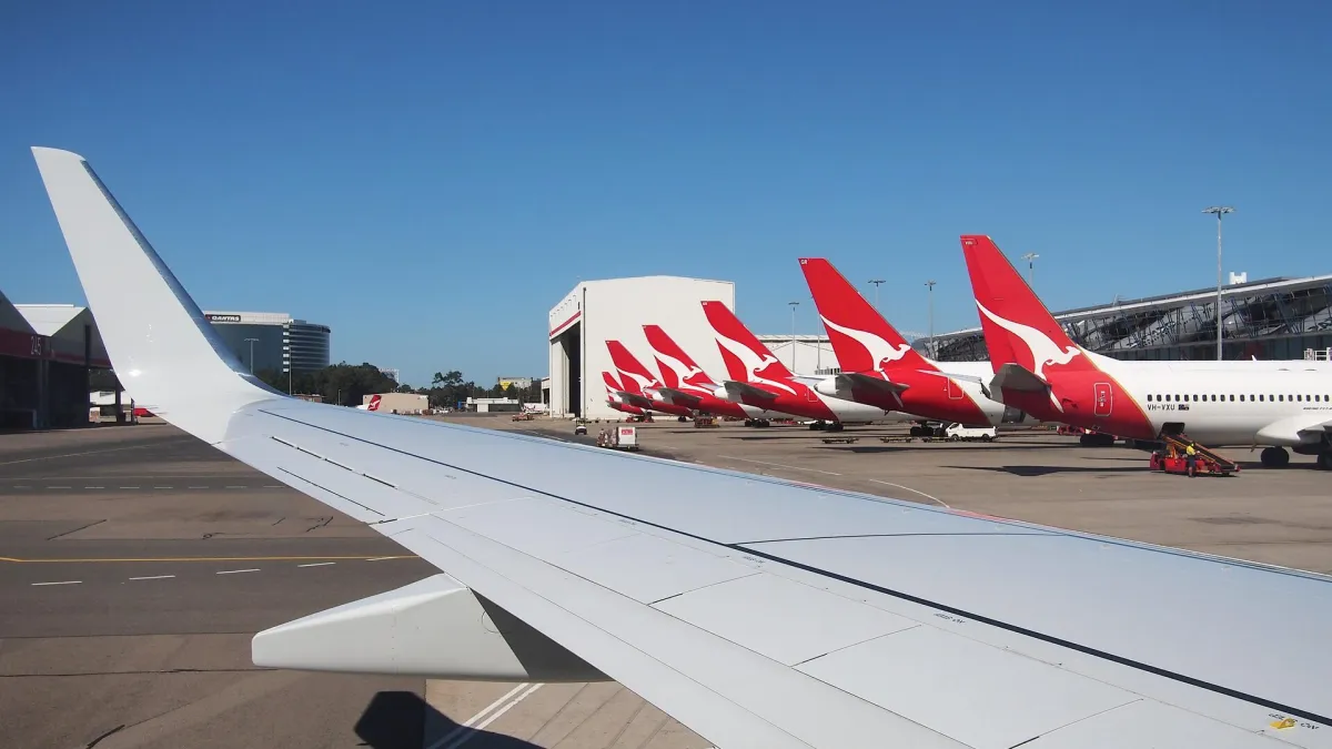 A line of Qantas aircraft with red kangaroo tails parked at Sydney Airport Terminal 3 under a clear blue sky, seen past an aircraft wing